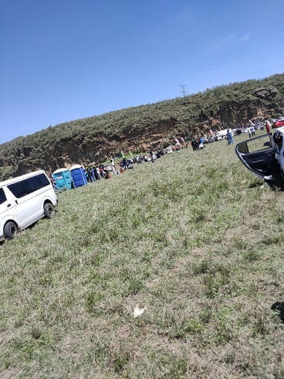 This image captures an outdoor gathering in the Olkaria ward, Kenya, under a clear blue sky. The scene is set on a large, uneven grassy field with patches of bare earth. In the foreground, a white van with off-road tires is parked on the left, angled towards the right. The open passenger-side door of a white car is visible on the far right. A single piece of white litter rests on the ground in the immediate foreground.
In the midground, approximately 30-50 individuals are gathered, interspersed among numerous parked vehicles, including cars and motorcycles. Two portable chemical toilets, one light blue and one dark blue, are visible near the white van. Several vertical flags or banners are also present within the gathering area. People appear to be standing, observing, or congregating; specific interactions are not discernible. One individual on the right side of the crowd is wearing a distinctive blue and white patterned outfit.
The background features a steep, rugged hill or cliff face densely covered with green and brown vegetation, consistent with dry scrubland. A power line pylon is faintly visible on the crest of this hill towards the right side.