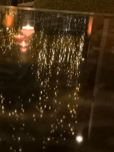 The image captures a serene nighttime scene featuring two floating candles on a dark, highly reflective surface, likely water. One candle is red and the other is pink, both emitting a soft glow, with their reflections clearly visible beneath them. The surface is adorned with countless small, bright, shimmering reflections, resembling scattered fairy lights or bokeh, creating a magical and ambient atmosphere. These lights appear to be reflections of decorative lights from the surrounding environment.

In the background, blurred vertical structures, possibly railings or window frames, and a fuzzy, textured object at the top (perhaps part of a sofa or upholstered seating) are dimly discernible. The overall impression is one of tranquility and romance, suggestive of a restaurant, hotel lounge, or a private residence in Palaio Faliro, Greece. The time of day is clearly night or very low light. No people or visible text are present.