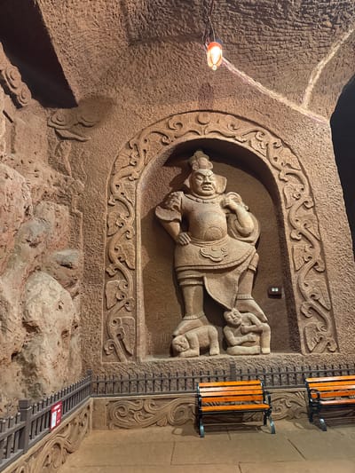 The image presents an intricately carved stone relief, possibly within the Longyou Caves, situated in Shizhong, China. The relief depicts a reclining male figure, clad in detailed armor or robes, positioned within an arched niche adorned with ornate floral and geometric carvings. The figure appears to be resting or sleeping, with two smaller figures shown in the background. The carving is illuminated by a warm light source on the left side of the image. A dark switch appears on the wall next to the top right of the carving. A section of ladder with an orange guard rail runs down the wall next to the carving. The surrounding area is a rough, textured stone, characteristic of the cave environment, indicating the relief is part of the larger cave structure. The image has been rotated so the view of the carving is horizontal.