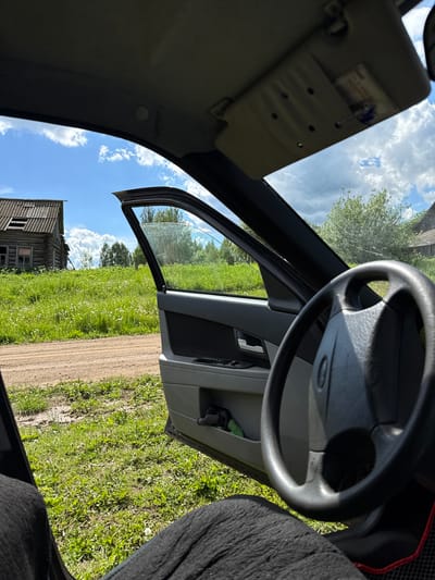 The scene is a view from inside a car, with the driver's side door open. Outside, a dirt road runs alongside a grassy field with white wildflowers. An old, weathered wooden building with broken windows stands in the background. The sky is blue with scattered clouds. The car's steering wheel is visible, and part of the dashboard and door panel can be seen. The location is likely Kamennyi Kliuch, Russia, based on provided context. There are no people visible in the image.