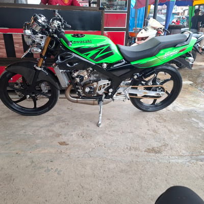A modified Kawasaki Ninja motorcycle is parked on a concrete surface in Rhieng Blang, Indonesia. The motorcycle is predominantly green with black accents and features carbon fiber parts on the front fender and fairings. It has chrome handlebars, a black seat, and a gold chain. The motorcycle is equipped with aftermarket exhaust pipes and a prominent radiator. In the background, there are other vehicles, including scooters, and a stall with red and blue awnings. A person wearing a red shirt is partially visible in the upper left background. The environment appears to be an outdoor marketplace or roadside stall area, with some wet patches on the concrete, suggesting recent rain.