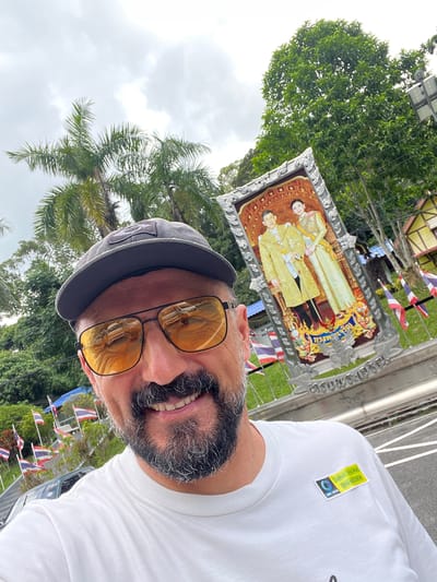 The image shows a man with a beard, wearing sunglasses, a cap, and a white t-shirt. He is smiling and appears to be outdoors. In the background, there is a framed picture of two people in formal attire, possibly royalty. There are also Thai flags visible, suggesting a patriotic or celebratory event. The setting appears to be tropical with palm trees and lush greenery, consistent with a location in Thailand. The sky is overcast, indicating a cloudy day. On the man's t-shirt, there is a yellow sticker with text that reads "Samui Viva Run" and a phone number "096-6928396." There are also some indiscernible symbols on the sticker. The overall mood of the image is positive and cheerful, with the man's smile and the festive decorations.