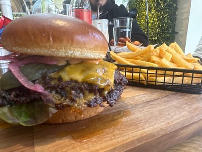 The scene depicts a meal served on a wooden cutting board in a restaurant located in Weißenburg in Bayern, Germany. A large double-patty smash burger, topped with melted yellow cheese, pickled red onions, dill pickles, and green lettuce, occupies the left side of the frame. To its right, a black wire mesh basket contains a quantity of golden-brown french fries. In the background, a person wearing a dark hoodie is partially visible, seated at the table, with a clear glass branded "granni" and a red glass bottle in front of them. A portion of a wall covered in artificial green foliage is visible behind the diner.