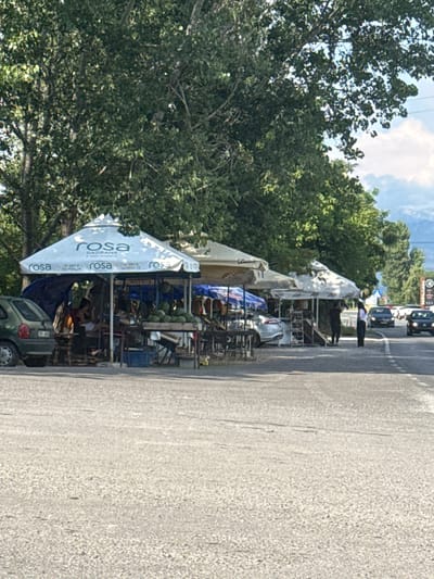 A low-angle, vertical shot shows a roadside fruit stand in Goričani, Montenegro. The stand is set up under large white umbrellas, one of which is prominently branded with the word "rosa" in blue lettering, repeated multiple times. Beneath the umbrellas, various items are displayed on shelves and tables, including what appear to be watermelons, bottled drinks, and other produce. Several people are visible behind the stand, seemingly tending to the goods or interacting with customers.
To the left of the stand, a dense canopy of green trees obscures the sky, with glimpses of white clouds and blue sky visible through the leaves. To the right of the stand, a paved road runs alongside, with cars and a few people standing on the sidewalk. One person in a white shirt and dark pants stands near the edge of the road, looking towards the stand. The road surface is a light grey asphalt, showing some texture and a few faint white lines.
The scene appears to be set during daylight, likely late morning or early afternoon, given the brightness of the sunlight and the shadows cast. The weather seems fair and sunny, typical for a summer day in the region. The overall impression is one of a bustling roadside market, offering fresh produce and refreshments to travelers. The presence of the umbrellas suggests efforts to provide shade from the sun. The "rosa" text on the umbrella indicates a possible brand affiliation, perhaps with a beverage company.