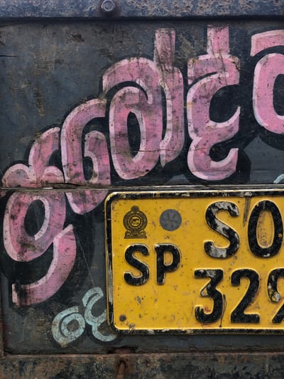 This media file is a close-up shot of a weathered, rustic surface featuring graffiti and a yellow license plate. The background appears to be a dark, possibly metal or wooden surface, marked with cracks and dirt. Large, stylized letters in pink and white graffiti are prominently displayed, some partially obscured.
To the right, a yellow license plate is affixed to the surface. The plate has raised black characters. Visible text on the license plate includes "SP 32" followed by other partially visible characters. There is also a circular emblem with an elephant image and decorative patterns on the plate. The surface of the license plate is scuffed and dirty, with visible wear.
On the lower right, a small section of pale blue graffiti, appearing to be numbers or letters, is visible. A metal hinge or bracket is also visible on the far right edge of the image, suggesting the surface might be part of a door, gate, or vehicle.
The overall impression is one of urban decay or a found object with layered markings. There are no visible people or animals. The lighting suggests it's daytime, but without strong shadows, it's difficult to determine the exact time or weather. The location context of Kodagoda, Sri Lanka, suggests this could be a piece of street art or signage from that region.