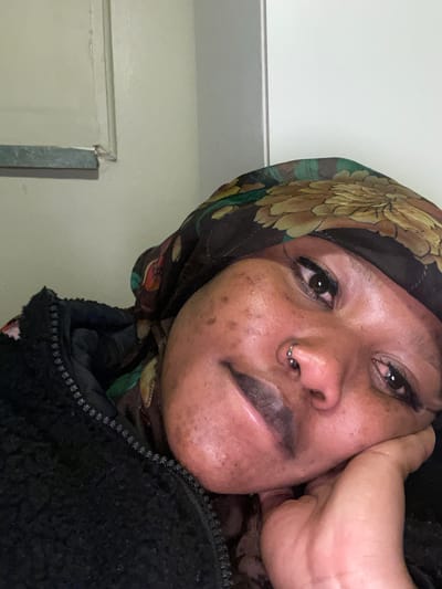 A close-up, angled selfie shows a Black female with light brown skin, freckles, and a nose ring resting her right cheek on her hand. She is wearing a black zip-up jacket and a headscarf with a floral print. The background appears to be a light-colored wall with a white doorframe to the right and a light-colored shelf or ledge to the left. The image was taken indoors, likely in a residential setting in Randburg, South Africa.
