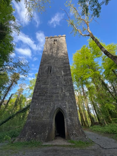 A low-angle, landscape-oriented photograph shows a tall stone tower in Knockaunbaun, Ireland, with surrounding trees and a blue sky. The tower, constructed from dark grey stones, dominates the frame. The top of the tower features crenellations. Two small windows punctuate the side of the tower. An arched entrance leads into the tower. The structure is situated amidst a forest, with green trees rising on both sides. The sky is a vibrant blue, punctuated by scattered white clouds and bare branches of trees. A paved area surrounds the base of the tower.