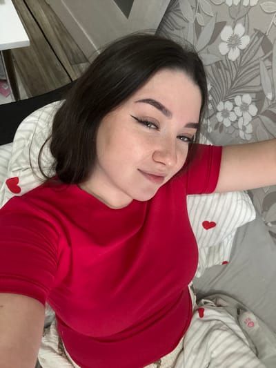 A young woman with long, dark hair and dark eyebrows is the primary subject of this image, captured in an overhead selfie perspective. She is lying down, looking directly at the camera with a relaxed expression and a slight, warm smile. Her eyes feature prominent black winged eyeliner.
She is wearing a plain red long-sleeved top or dress. Her right arm is raised, with her hand likely holding the camera. The woman is positioned on a bed or soft surface. Behind her head is a white pillow with subtle gray stripes and scattered red heart motifs. To her right, another striped fabric with red hearts is visible, possibly part of the bedding or another pillow. Further in the background, a plain gray pillow or blanket can be seen, along with a glimpse of a light gray soft object that has the pink number "50" printed on it.
The background on the left side of the image features wallpaper with a floral pattern, showing white flowers and gray leaves against a lighter gray background. Below the wallpaper, on the far left, there's a small section of what appears to be wooden flooring or wall paneling.
The setting is an indoor, domestic environment, likely a bedroom, situated in Башкултаево, Russia. The lighting suggests it is daytime. The overall mood conveyed by the woman's expression is pleasant and content.