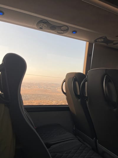 The image captures a view from inside a moving bus or coach, looking out of a large side window during either sunrise or sunset. The interior shows the upper portion of a dark gray or black passenger seat with quilted stitching in the immediate foreground to the left. To the right, two empty, forward-facing dark gray seats with integrated grab handles on their backs are visible. Above the window, a light gray overhead panel features ventilation outlets, passenger service buttons, and two illuminated blue LED lights.

The exterior view reveals a wide, slightly hazy landscape under a sky transitioning from pale blue at the top to a warm, yellowish-orange at the horizon. The terrain below is predominantly a reddish-brown color, dotted with scattered dark vegetation, possibly olive groves. In the mid-distance, a distinct cluster of buildings indicates a settlement, identified as the city of Carmel, Palestinian Territory, from an elevated perspective. A thin, dark horizontal line, resembling a power line, crosses the upper mid-ground of the exterior view. No individuals are visible within the frame.