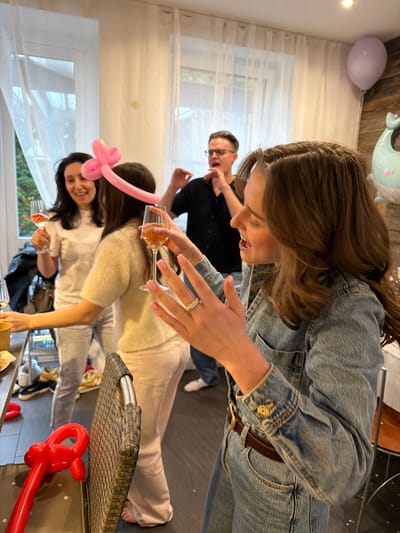 The image shows a group of people indoors, apparently celebrating. A woman in a denim jacket holds a champagne flute. A man with glasses stands in the background. Another woman, wearing a pink balloon headband, smiles and also holds a glass. The scene includes balloons, indicating a celebratory event. The location is Крючково-2 Снт, Russia.