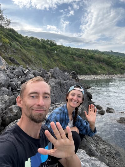 Two people, a man and a woman, are posing for a selfie on a rocky outcrop next to a body of water. The man is in the foreground, wearing a black t-shirt with a blue design and has a short beard. The woman is behind him, wearing a blue plaid shirt and a light blue cap, and she is smiling. Both are waving. The background features a hillside covered in green vegetation, a body of water, and a cloudy sky. The image does not contain enough information to identify the location as Bondarenkovo, Ukraine.