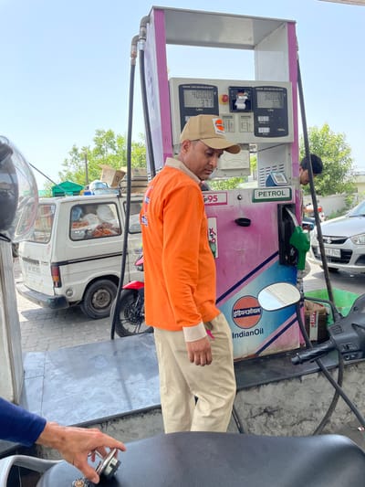The image shows a man in an orange shirt and khaki pants standing at a gas pump at an Indian Oil petrol station. The pump is pink and blue with the "Indian Oil" logo prominently displayed. The pump display shows "400.00" and "3.71 Litre" and "300.00" and "3.03 Litre". The gas nozzle is green. Behind the man, there is a white Omni van with the license plate "RJ 39 U092". A black motorbike is parked behind the van. In the background, a white car with the license plate "RJ 09 F 7029" can be seen. The location is Sidhpur, India.