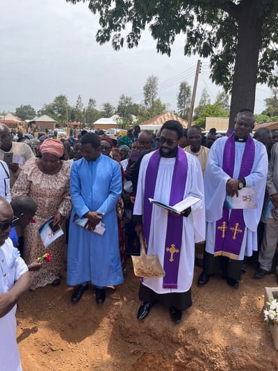 A solemn outdoor gathering is underway in Dogon Dinia, Nigeria. Two men, possibly clergy, stand in white robes with purple stoles marked with gold crosses, each holding a book or document. Numerous attendees, including women in patterned dresses and headwraps and men in a mix of clothing, surround them. One woman holds a red rose. The setting appears to be a gravesite, evidenced by the dirt ground, and a possible headstone with white flowers is visible in the background. There is an electric utility pole. The sky is overcast.