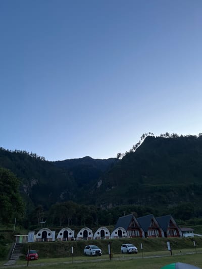 The image depicts a serene evening scene in Merodot, Indonesia. The sky is a clear, gradient blue, transitioning to a lighter hue towards the horizon. In the foreground, a row of unique dome-shaped cabins and three triangular A-frame houses are situated on a grassy slope. A red car is parked to the left of the cabins, and a white SUV is parked in front of the A-frame houses. The middle ground is characterized by lush green grass and several trees. The background is dominated by a majestic, dark green mountain range covered in dense forestation. The mountain slopes are steep, with scattered trees silhouetted against the sky along the ridgeline. The overall atmosphere is peaceful and tranquil, suggesting a secluded natural retreat.