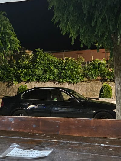 The image shows a dark-colored sedan parked in front of a stone wall with dense green bushes behind it. A large tree trunk is visible on the right side of the frame. In the foreground, a weathered wooden surface with some papers on it is present. The scene appears to be outdoors at night, with artificial lighting illuminating the car and vegetation. The context of Biaga, Bulgaria is not directly discernible from the visual elements in the image itself.