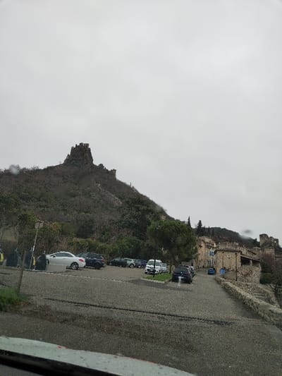 The image depicts an overcast day in Rochemaure, France, showing a village at the base of a prominent, rugged hill topped with castle ruins. The foreground presents a wet, paved surface, indicative of a parking lot or road, possibly viewed through a vehicle's windshield due to visible streaks and reflections.

In the mid-ground, a parking area contains multiple vehicles: a white sedan, several dark-colored SUVs and sedans, and a white utility van. Several dark streetlights and waste bins are also present. To the right, traditional stone buildings, characteristic of a European village, line a curving road that descends into the scene. These structures feature visible stone masonry, tiled roofs, and some with pale blue or grey painted facades. A stone retaining wall with steps is visible on the far right.

The background is dominated by a steep, treeless hill, sparsely covered with dark, bare deciduous vegetation, suggesting a cold season. At its summit and along its upper slopes are substantial stone ruins, consistent with a medieval fortress or castle. To the far right, additional hills or mountains are partially obscured by mist or low clouds. The sky above is uniformly grey and overcast. No individuals are visible.