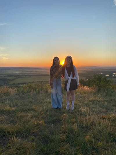 Two young females stand on a grassy hilltop overlooking a valley bathed in the warm glow of a sunset. The female on the left wears a plaid shirt tied at the waist and light-wash flared jeans. The female on the right has a white headband, a dark dress, and a white cardigan tied around her shoulders. They are positioned side-by-side, facing the camera, with the setting sun directly behind them, creating a silhouette effect. The background depicts a vast, green landscape with hints of water bodies and distant trees, characteristic of the rural scenery found near the village of Uzyytamak, Russia. The sky transitions from a clear blue at the top to vibrant orange and yellow hues near the horizon where the sun is setting. The foreground is covered in dry, textured grass, illuminated by the golden hour light.