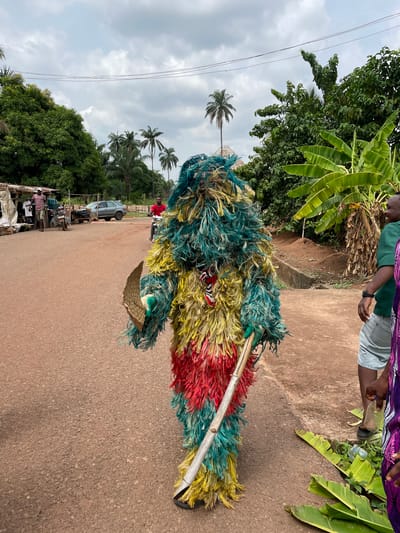 A masquerade figure, predominantly composed of vibrant blue, yellow, and red synthetic fibers, is seen on an asphalt road in Agba Umana, Nigeria. The masquerade holds a small, woven basket and a long, smooth pole. An individual in a green shirt and denim shorts stands at the edge of the road next to banana trees. Behind the masquerade, a gray sedan is parked near market stalls. Palm trees rise in the background under a cloudy sky.