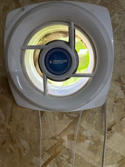 The image shows a white circular exhaust fan mounted on a wall constructed from oriented strand board (OSB). The fan has a central blue circular logo with white stars and text that reads "SPARK LUX professional tools." The fan appears to be partially open, revealing a brightly lit outdoor scene with green foliage visible through the opening. Two white electrical cords hang down from the fan. The overall impression is of an interior space, possibly a workshop or shed, with an exterior view visible through the fan. The specific location in the city of Айет, Kazakhstan, cannot be determined from the visual information provided in the image.