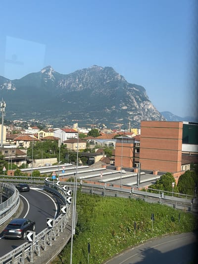 A slightly rotated eye-level shot reveals a detailed view of the city of Lecco, Italy, situated at the foot of a mountain range under a clear, bright sky. The cityscape features residential and commercial buildings, a highway overpass with black cars and road signs, and a vibrant green lawn and roadway. The mountain looms as a backdrop, adding depth to the scene. The architectural elements of Lecco are prominently displayed.