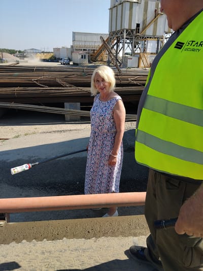 An outdoor, daytime, medium shot captures a woman in a floral dress standing in front of a large pile of steel rebar at a construction or industrial site in Svityn, Ukraine. She is holding a long pole with a bottle attached to its end. To her right, a person wearing a bright yellow safety vest with "STAR SECURITY" printed on it partially obstructs the view. In the background, industrial buildings, stacks of rebar, and a vehicle are visible under a clear blue sky. The ground is a mix of asphalt and dirt.