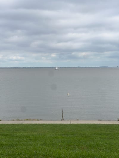 The image depicts a calm, overcast day at a body of water, likely a bay or sea, near Wilhelmshaven, Germany. In the distance, a solitary sailboat with white sails navigates the water. Along the horizon, wind turbines are visible, suggesting an industrial or coastal area. Closer to the foreground, a white buoy floats on the surface. A concrete embankment lines the water's edge, and beyond that, a grassy slope descends towards the viewer. The sky is filled with heavy, grey clouds. There are no people visible in the image.