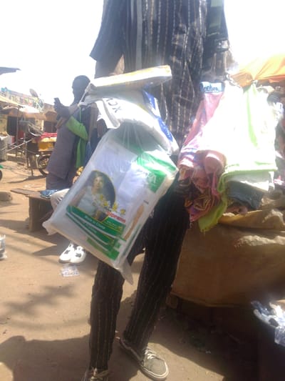 An outdoor market scene in Mista Ali, Nigeria, is depicted under bright sunlight. In the immediate foreground, the lower torso and legs of an individual are visible, clad in a dark, vertically striped shirt and trousers, with grey canvas shoes on their feet. This individual is carrying multiple items, including a clear plastic bag containing a white package labeled "Supreme" with Chinese characters and "Made in China," featuring an image of a child, likely diapers or similar goods. Adjacent to this, a bundle of colorful fabrics, predominantly pink, green, and blue, is also carried. To the left of the foreground subject, a second individual is seated on a low wooden bench, dressed in a light-colored robe with a green overlay, actively holding and looking down at a smartphone with both hands. A pair of white shoes with black details rests on the dusty ground near this seated person. The background reveals a bustling market environment with indistinct figures, temporary structures, and stalls with fabric coverings under a bright, hazy sky. Shadows are cast on the dry, unpaved ground.