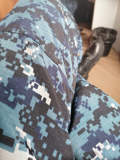 The image shows a close-up view of camouflage fabric, likely part of a uniform or clothing. The pattern consists of pixelated shapes in shades of blue, black, and white. In the background, out of focus, a person is wearing a black boot and a dark object, possibly a trash can, is visible. The setting appears to be indoors with a wooden floor. The image does not provide any information about the city of Ust'-Chem, Russia, or any specific location within it.