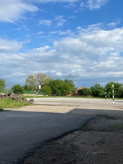 The image shows an outdoor scene, likely in a rural or semi-rural area. The sky is blue with numerous white clouds. On the right side of the image is a paved road with some wear and tear. To the side of the road, there is a grassy area with trees and a fence. In the background, a building with a red tile roof can be seen. There are also some black and white barriers on the paved area. Based on the context, the location is likely Rakevo, Bulgaria. The image does not contain any people. There is a basketball hoop. There are some old tree logs next to a green fence.