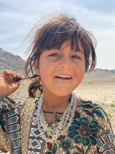 A young girl stands outdoors in what appears to be a rural or arid area. She has brown hair, brown eyes, and is smiling. She is wearing a patterned dress with large floral designs and geometric shapes, along with a multi-strand clear beaded necklace. The background includes a barren landscape and a clear sky. The girl is holding a strand of her hair in her right hand. The location is possibly near the city of Ashoqah in Afghanistan.