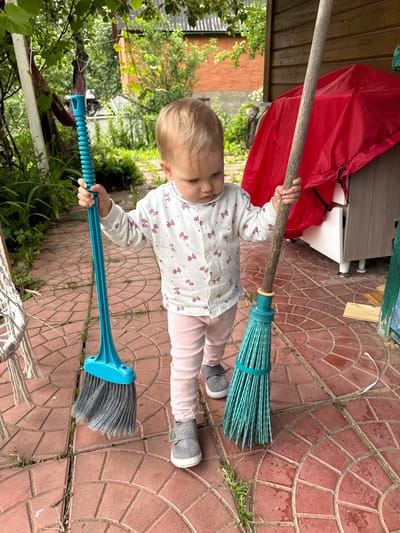 A young child, with blonde hair and fair skin, is shown from the chest down, holding two brooms. The child is wearing a white long-sleeved shirt with a repeating pink bow pattern and pink pants. Gray sneakers are on the child's feet. The child is positioned on a patio made of red, circular patterned bricks. In the background, a red tarp is visible, as well as green foliage and wooden structures, suggesting an outdoor setting. The overall lighting implies it is daytime. The child's posture and the way they are holding the brooms suggest they are engaged in play. One broom is positioned above the child's head and appears to be a traditional broom with a wooden handle and teal bristles. The second broom is held lower, closer to the ground, with a blue plastic handle and gray bristles. The image is oriented vertically, with the child and brooms appearing in the center. The scene suggests a casual, domestic outdoor environment, possibly a backyard or garden area.