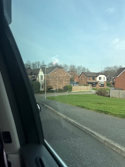 The image displays a residential street in Bury, United Kingdom, observed from within a vehicle through its right-side window. The left portion of the frame is occupied by the vehicle's dark door panel and window frame. The foreground shows a dark grey tarmac road, delineated by a light grey concrete curb. Beyond the curb, a strip of vibrant green grass extends. The midground features a row of modern detached and semi-detached houses. These houses are primarily constructed of reddish-brown brick with dark grey tiled roofs. One prominent house on the left is lighter-colored, possibly rendered white or cream, featuring a bay window and a dark green manicured hedge. A dark grey street light pole stands near the curb in front of this house. Further right, an open grassy area is visible, bordered by wooden panel fences. The background consists of a dense line of bare deciduous trees, indicating a winter or early spring season. The sky is clear and light blue, exhibiting a distinct white, helical vapor trail or smoke plume in the upper-mid section. No people, animals, or active interactions are visible.
