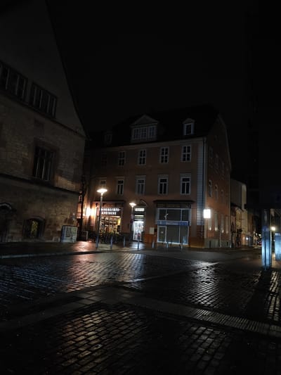 The image depicts a wet, cobblestone street scene at night in Göttingen, Germany. The "Universitäts-Apotheke" (University Pharmacy) is prominently visible with brightly lit signage. The pharmacy and adjacent buildings are illuminated by streetlights, reflecting off the wet pavement. Several bicycles are parked near the pharmacy. Other buildings are visible in the background, with a few additional lights suggesting a street extending into the distance. The overall atmosphere is dark, emphasizing the contrast between the lit buildings and the unlit street. There are no people visible in the image.