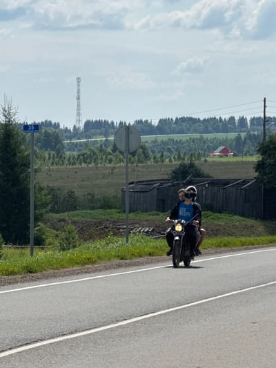 A rural roadside scene features a motorcyclist and a passenger riding a motorcycle on a paved road with white lane markers. The motorcyclist is wearing a black helmet and a blue t-shirt with text that appears to say "VSE KRUTO". The passenger is wearing a white helmet. In the background, there are several dilapidated wooden structures, a green field, a line of trees, and a red and yellow building. A cell tower is visible in the distance. A blue street sign with white text reading "35" and "ПЕРМСКИИ КРАЙ" is posted on a pole to the left of the road. The sky is partly cloudy.