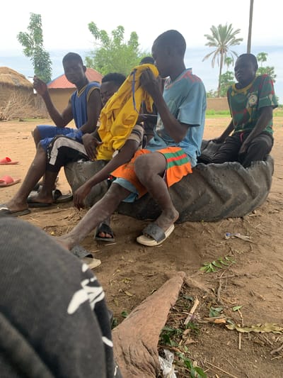 The media file shows a group of young boys gathered outdoors in a rural setting, likely Gwolong, Nigeria. The scene appears to be during the daytime, with bright natural light suggesting fair weather. The ground is dusty and dirt-covered, with scattered dry leaves and twigs, hinting at a dry season. In the background, a palm tree is visible, along with some thatched-roof buildings and lush green foliage, indicating a tropical or sub-tropical environment.
The central focus of the image is a large, worn tire, around which several boys are interacting. One boy, wearing a blue and yellow jersey, is sitting on the tire, holding what appears to be a cigarette. Another boy, in an orange and green striped shirt, is also positioned near the tire, with his legs bare and dusty. A third boy in a blue t-shirt is partially visible. The boys are dressed in casual, everyday clothing, some of them appearing to be made of light fabrics suitable for a warm climate. Their skin is dusty, consistent with the dry, outdoor environment.
The activity seems informal, with the boys relaxing and socializing around the tire. There's a sense of camaraderie and possibly boredom or idleness. The overall mood is casual and somewhat somber due to the boy holding the cigarette, but the bright lighting and presence of children suggest a typical day in the community. No visible text is present in the image.