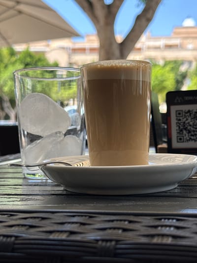 A close-up, eye-level shot shows a table outdoors with two glasses and a QR code visible in the background. On the right, a clear glass contains a light brown beverage with foam on top, placed on a white saucer with a spoon. To the left, another clear glass is filled with ice cubes. The table's surface is a dark, woven material. In the background, blurred green foliage and a building suggest an outdoor cafe or restaurant setting in Campillo Del Moro, Spain, under a bright sky.