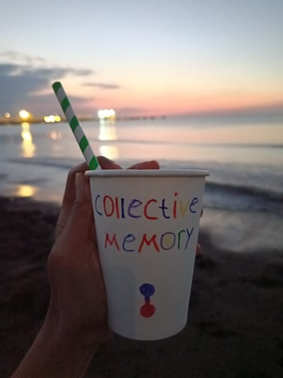 A person's hand holds a white paper cup with a green and white striped straw. The cup has colorful handwriting on it. The top line reads "COLLECTIVE" in red, blue, and yellow. The second line reads "MEMORY" in red, yellow, and blue. Below the text, there is a small drawing of two colored circles, one red and one blue, connected by a thin line. The background shows a beach scene at dusk. The sky has a gradient of colors, from pale blue at the top to pink and orange near the horizon. The ocean is calm, with gentle waves reflecting the lights from distant structures on the shore. The sand in the foreground is dark and textured. The overall mood is peaceful and contemplative, possibly evoking a sense of nostalgia or shared experience, given the text on the cup. The lighting suggests it is either sunrise or sunset.