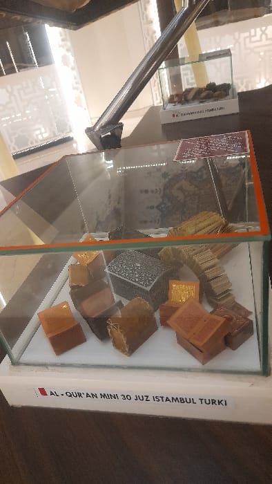The image captures an indoor display within a museum or exhibition space located in Glantengan, Indonesia. The primary subject is a large rectangular glass display case resting on a white base. Inside, numerous miniature Qur'ans are exhibited. These miniature books vary in appearance, with some featuring brown covers, others a metallic gold finish, and one prominent, intricately patterned dark silver-colored case or book. Several books are displayed open, revealing their internal pages. A notable arrangement includes a large arc constructed from a stack of miniature books. A white label affixed to the base of the main display case reads "AL-QUR'AN MINI 30 JUZ ISTAMBUL TURKI," indicating the origin and content of the miniatures.
In the background, a smaller glass display case is visible, containing several dark, irregular, possibly ancient artifacts. The general environment features light-colored walls or screens with decorative geometric patterns, consistent with Islamic architectural motifs. A reflective, dark-colored floor is present in the foreground and extends into the midground. A polished metal structural beam or support column is prominently positioned in the upper-left and center of the frame, reflecting ambient light. No individuals are present in the scene.