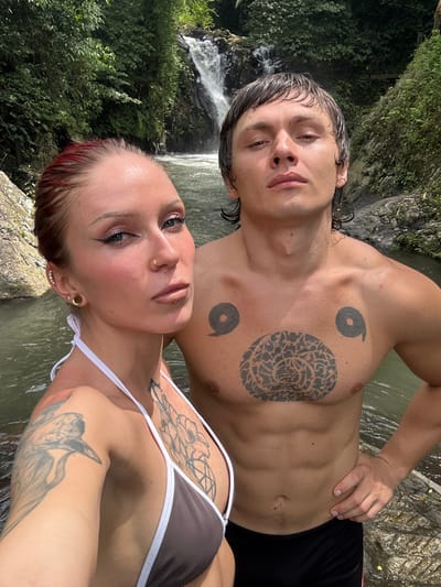 Two individuals, one female and one male, are depicted in a selfie-style photograph standing in shallow water in Ambengan, Indonesia, with a waterfall and dense tropical foliage in the background.
The female subject is positioned closer to the camera on the left. She has red hair, light skin, and is wearing a brown bikini top with white trim. Visible facial piercings include a gold-colored nose ring in her left nostril and a labret piercing below her lower lip. Her left ear features multiple piercings, including a large gold-colored ear tunnel and smaller studs. Tattoos are visible on her left shoulder/upper arm (depicting a bird) and the upper left side of her chest (a floral or plant design). She looks directly at the camera with a neutral expression.
The male subject stands slightly behind and to the right of the female. He has short, dark, wet hair and light skin, and is bare-chested, wearing black swim shorts. Three black tattoos are visible on his chest: two circular, stylized patterns on each pectoral muscle and a large, intricate circular design at the center of his chest. His left hand rests on his hip. He also looks directly at the camera with a neutral expression.
Behind the individuals, a multi-tiered waterfall cascades into the body of water they are standing in. The surrounding area is characterized by lush green tropical vegetation and visible rock formations along the water's edge and within the stream. The water appears clear to light brown. The scene is brightly illuminated by daylight.