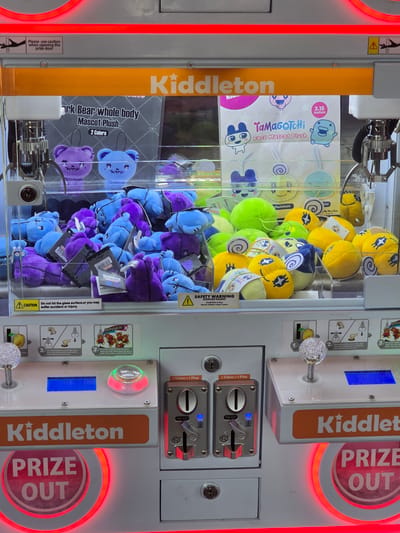 The image shows a Kiddleton prize claw machine filled with plush toys. The toys appear to be small, stuffed bears in purple and blue, and spherical plush toys in yellow and green. Some of the toys have tags with Japanese writing. The machine has two joysticks and coin slots. The overall setting is an indoor entertainment area, likely an arcade or mall. There are no people visible in the image. The location is specified as Cumming, United States of America.