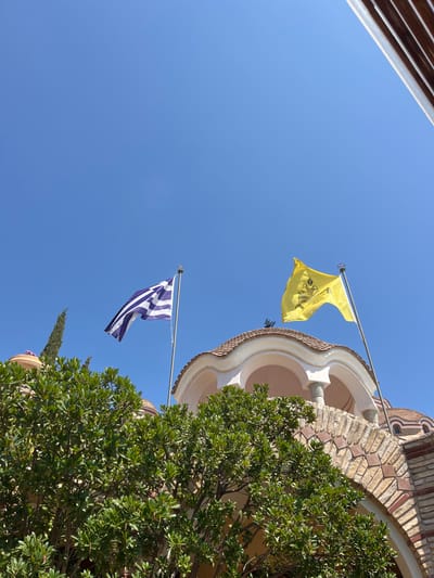 The image shows a low-angle view of the Moni Archaggelou, a monastery located in the city of Moni Archaggelou, Greece. The image is captured from within the monastery grounds, looking upward towards the sky. The scene is dominated by a clear blue sky and two flags fluttering in the wind.  The Greek flag is visible on the left and a yellow flag with an emblem on the right. Both flags are flying from tall flagpoles against a backdrop of a clear blue sky.  A tall green tree is located in the bottom left of the image. The main structure of the monastery appears in the background, characterized by a white-trimmed, domed building with a partially visible archway. The archway is constructed of red brick. The structure is topped with a white roof, hinting at the architectural style of the monastery.  Green foliage partially obscures the structure, suggesting a lush and verdant environment. The overall scene depicts a tranquil and serene setting, possibly a popular pilgrimage site.