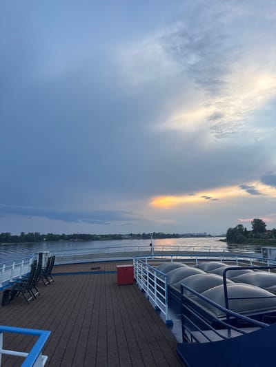 The image captures a view from the deck of a ship on a river, possibly the Nizhny Novgorod Reservoir, near the city of Imeni Sverdlova, Russia. The sky is a mix of dramatic grey clouds and a bright, sunlit band of orange and yellow. The river is calm, reflecting the sky, and a distant shoreline with trees and buildings is visible. On the ship's deck, there are several empty lounge chairs arranged on a wooden deck, a red storage container, and a section with large, curved, translucent covers over what appears to be a lower deck or cargo area. A railing with white posts and blue trim separates the deck from the water. The overall atmosphere is serene, with the dynamic sky contrasting with the stillness of the water and the deck.