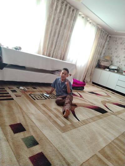 A young boy is sitting cross-legged on a patterned rug in a room. He is wearing a blue t-shirt and brown pants. He is looking at the camera and holding up two fingers in a peace sign. In front of him, there is a smartphone and a white power adapter with red and white cords. The room has white curtains, a white cabinet, and floral wallpaper. The scene appears to be in a residential setting.