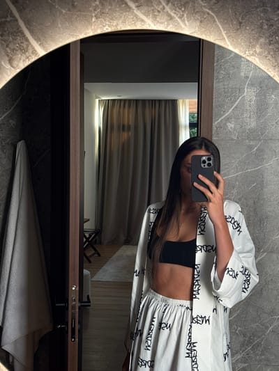 In an indoor setting, likely a hotel room or apartment in Bakovići, Montenegro, a young woman stands in front of a large, circular mirror with integrated lighting. She is taking a selfie with her smartphone, which is held in her right hand. The woman has long, dark hair and is wearing a black bralette and a matching white pajama set with black lettering. The outfit consists of a robe-like top and loose-fitting pants.
The background reflected in the mirror shows a room with light brown curtains, wooden flooring, and a small stool with a folding base. To the left of the woman, also reflected in the mirror, is a doorway with a wooden frame and a black door handle. A grey towel is hanging on a hook to the left of the doorway. The walls are decorated with a marbled pattern. The overall lighting is dim, with the mirror's light providing a primary source of illumination.