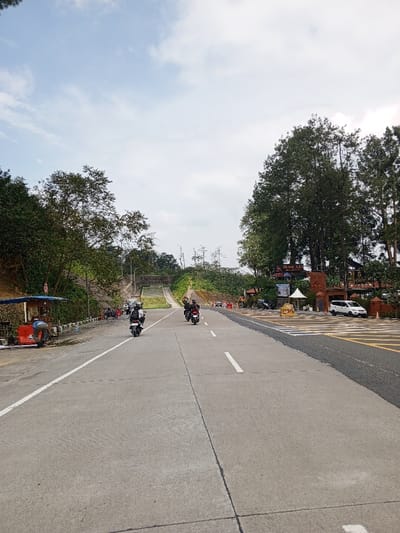 The image depicts a concrete two-lane road in Ciloto, Indonesia, viewed from a low perspective. The sky above is partially overcast with visible blue patches.
On the left side of the road, a blue-canopied roadside stall with a red base is present, attended by an individual in a dark shirt bending down. Behind the stall, a sloped embankment is covered with dense green trees and vegetation. A low concrete barrier separates the road from this area. Further down the road on the left, an overpass or bridge structure spans above the roadway.
On the road, at least three motorcycles are visible, traveling away from the viewer. One motorcycle is in the left lane, and two are in the right lane, with riders present. The road features white solid and dashed lane markings. To the right of the road, the surface transitions to a darker asphalt patch before reaching a complex entrance structure. This entrance, featuring "BUMI AKAR" signage, is constructed from reddish-brown materials and is flanked by tall, mature evergreen trees. A white car is parked in a paved area beyond this entrance. Small white tent-like structures and other architectural elements are visible around the entrance. The background on both sides of the road is dominated by dense green foliage on elevated terrain.