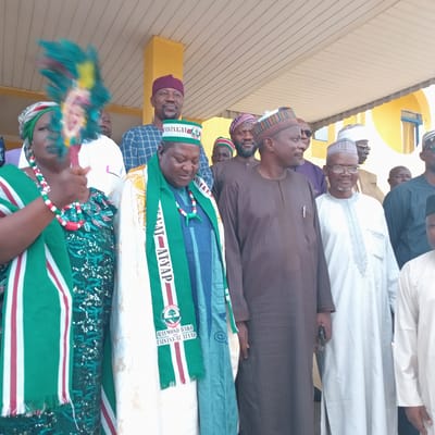 In Unguwar Gaya, Nigeria, a group of men and one woman are gathered outdoors, standing in front of a yellow building with a beige, slatted awning. The woman on the far left is holding a colorful fan, decorated with turquoise, yellow, and red feathers. She is wearing a green and red beaded necklace and a green outfit with intricate patterns. To her right, a man in a blue and white checkered shirt and a maroon cap is looking towards the camera. Next to him, a man wearing a green scarf with white and red stripes is visible. He is also wearing a cap. Further to the right, a man in a brown tunic and cap is looking to the side. Another man in a white cap and glasses is looking forward. The man to his right is wearing a dark blue outfit. The man furthest to the right is wearing a dark outfit and a black cap. The environment appears to be a public space, possibly for an event or gathering.