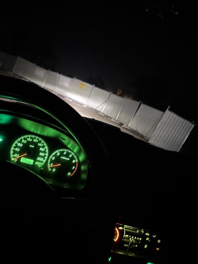 The image, captured at night in Suhache, Bulgaria, shows the interior perspective from a stationary vehicle. The car's dashboard, prominently displayed, features a speedometer indicating 0 km/h and a tachometer at 0 RPM, both illuminated in green. A digital trip meter reads "116.1". A dark steering wheel is partially visible in the foreground. To the lower right, the vehicle's climate control panel, illuminated with orange and green lights, shows the fan speed set to "OFF". Through the windshield, the vehicle's headlights illuminate a segment of ground directly ahead, leading to a long, light-colored temporary construction fence made of corrugated metal panels. Multiple yellow warning signs are affixed to this fence. The area beyond the fence is obscured by complete darkness. No individuals are visible in the scene.