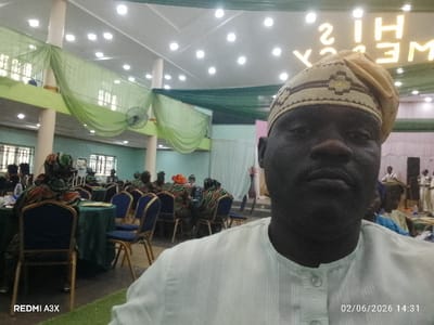 The image captures an indoor event, likely a social gathering or function, from the perspective of a person taking a selfie.

In the foreground, a man with a dark complexion is prominent, looking directly at the camera. He is wearing a light-colored, possibly white or cream-colored, traditional top with a patterned neckline and placket, and a matching or coordinated patterned cap (fìlà) with embroidered details. His face fills a significant portion of the lower right and center of the frame.

Behind him, a spacious hall is visible, set up for an event. Several round tables, covered with dark green tablecloths, are arranged throughout the room. Each table has multiple chairs with blue upholstered seats and light brown/gold frames. Many attendees are seated at these tables, some visible from the back, wearing various outfits, including what appear to be traditional patterned garments.

The hall itself is decorated with green drapes or fabric swags suspended from the ceiling and draped between pillars. The ceiling features numerous recessed light fixtures that are illuminated, providing ample light. One ceiling fan is clearly visible hanging from the ceiling. On the left side of the room, there are windows allowing natural light to enter.

A large, illuminated sign is prominently displayed on a back wall, spelling out "HIS MERCY." Due to the selfie camera capturing a mirror image, the text appears reversed.

In the bottom left corner of the image, the watermark "REDMI A3X" is visible, indicating the device used to capture the photo. In the bottom right corner, the date and time "02/06/2026 14:31" are stamped on the image. The overall atmosphere appears to be one of an organized gathering or celebration during the daytime.