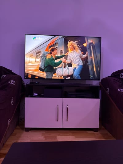 The image shows a modern living room with a large flat-screen television mounted on a lavender-colored wall. The television displays a scene from a film or show featuring two people. The person on the left is a woman with blonde, curly hair, wearing a striped shirt and blue jeans. She is holding onto a rope or strap with her hands extended upwards. The person on the right is a young man with curly brown hair, wearing a green hoodie and blue jeans. He is holding onto the same rope or strap, appearing to be pulled or lifted. Both individuals seem engaged in a physical activity.
Below the television is a sleek, minimalist entertainment unit with two white cabinet doors and a dark shelving unit. The shelving unit contains a black device, possibly a game console or media player, and some cables.
To the right of the entertainment unit, a portion of a dark brown couch is visible, with a dark purple patterned throw blanket draped over it. The lighting in the room is predominantly purple, suggesting ambient lighting or mood lighting.
The television screen displays the "Avito" logo in the lower left corner, which is a Russian classifieds and e-commerce website. There is also a "TBU" logo and "12+" age rating visible in the upper right portion of the screen. The overall scene suggests a casual, indoor setting, likely a home entertainment area. The time of day is not discernible.