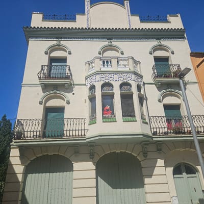 The image depicts a three-story building in La Jonquera, Spain, notable for its architectural details and use of color. The facade is primarily light beige with pale green accents on the trim, window frames, and arched doorways. Blue and white tiles are integrated into the design along the upper portion of the building and in the central bay window. The ground floor has three arched openings with large wooden doors. The second and third floors feature arched windows with metal balconies. Most of the windows have the pale green shutters closed. The most striking detail is a red Elmo puppet visible in the center window of the bay on the second floor. A light pole stands to the right of the building. The sky is clear and bright blue. A small green tree is visible to the left of the building.
